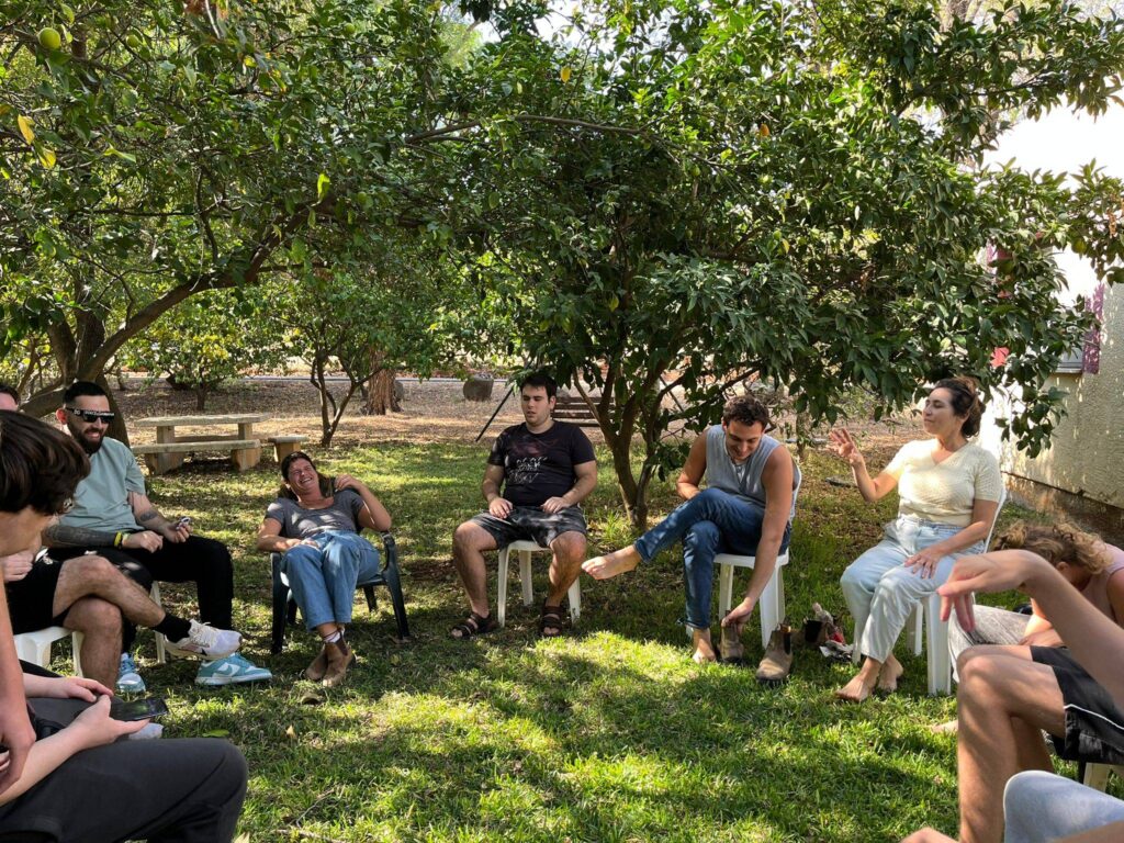 קבוצה יושבת במעגל מתחת לעצים בפארק, מדברים ומחייכים. אווירה נינוחה וירוקה.
