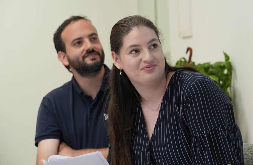 שני אנשים יושבים יחד בחדר מואר, מביטים לכיוון לא מוגדר, עם מסמכים בידיהם.