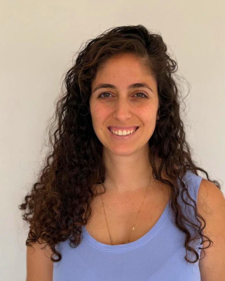 אישה מחייכת עם שיער מתולתל וארוך, לובשת חולצה סגולה בהירה, רקע לבן.