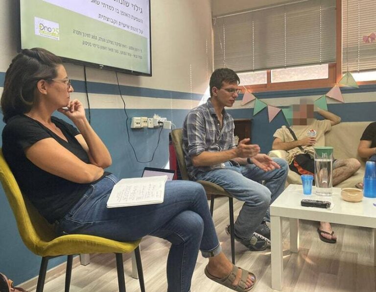 קבוצה בדיון בחדר עם מסך, שלטים צבעוניים על הקיר ושולחן עם כוסות מים.