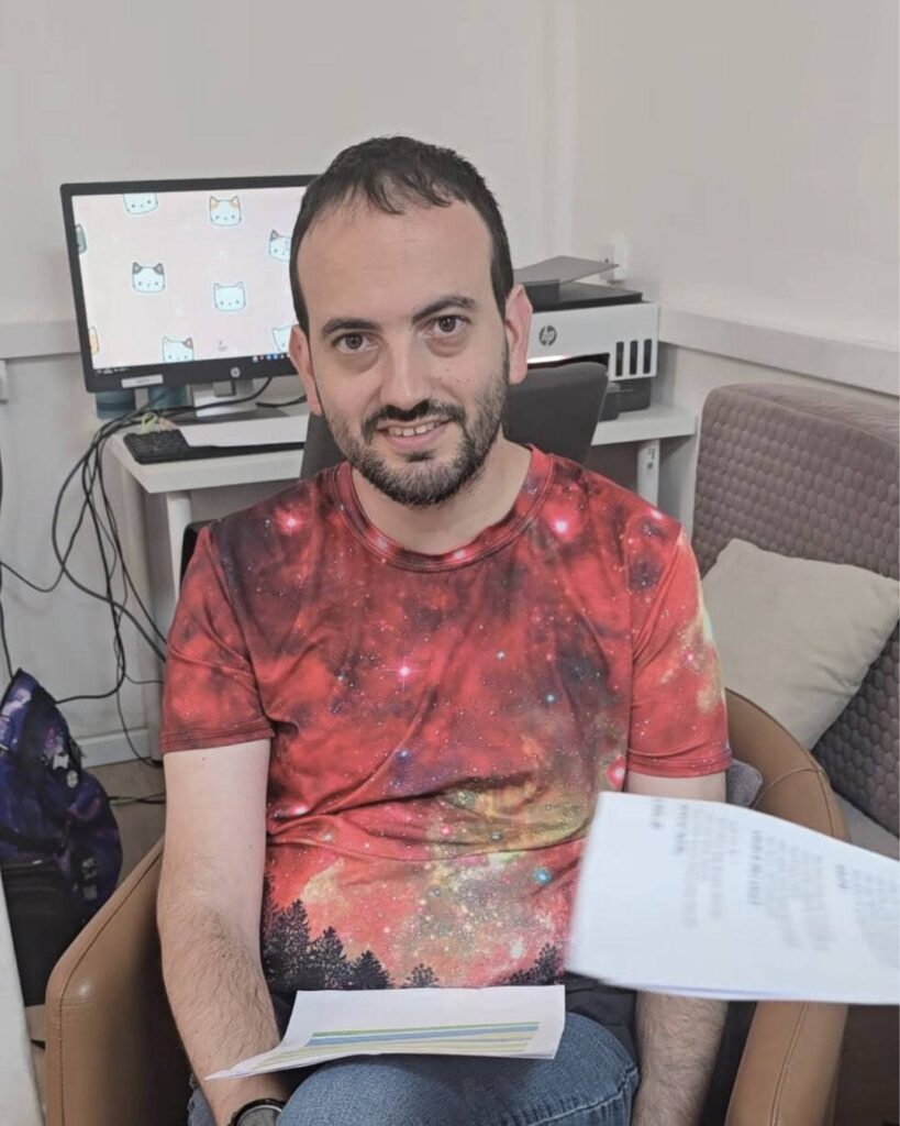 אדם יושב בכורסה, לובש חולצה צבעונית עם הדפס גלקסיה, ברקע מחשב עם תמונת רקע של חתולים.