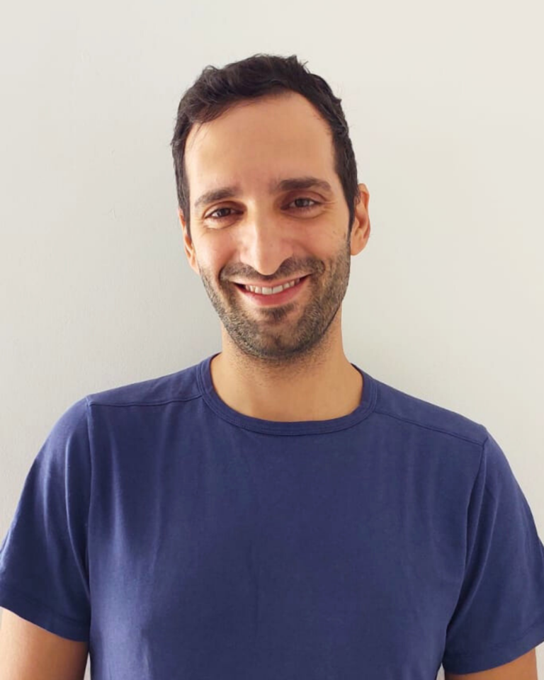 אדם עם חיוך וחולצה כחולה עומד מול קיר לבן.